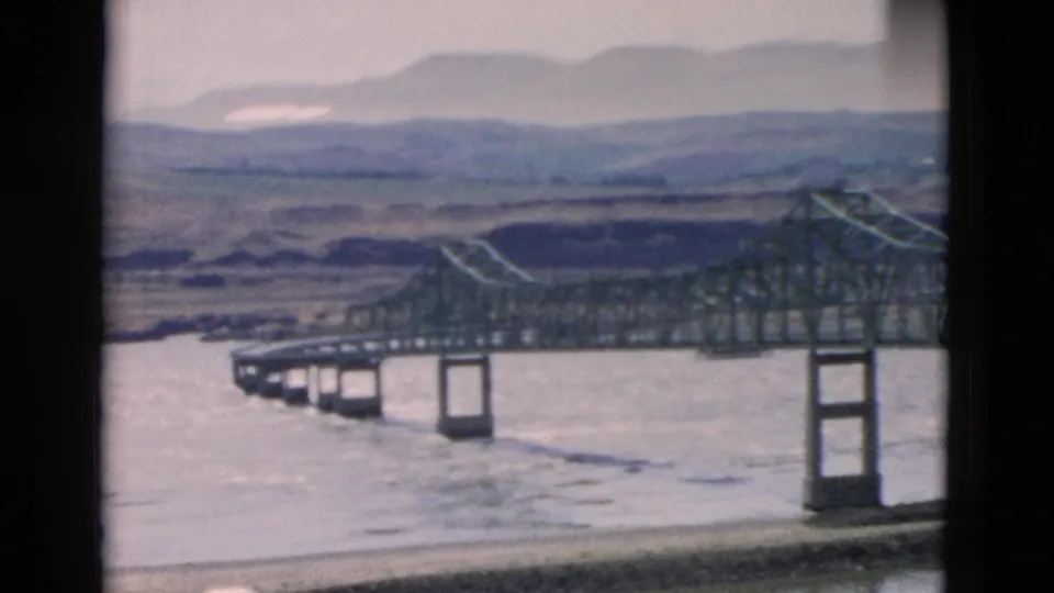 The pan reaches a long steel truss bridge on concrete piers over a broad river, with low hills and shoreline visible behind it.