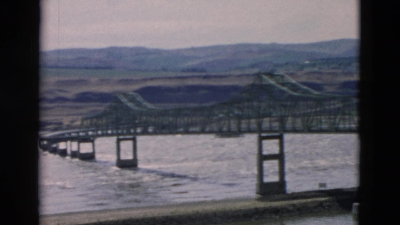 Vintage 8mm view of a steel truss bridge over a broad river, with concrete piers and distant rural hills.