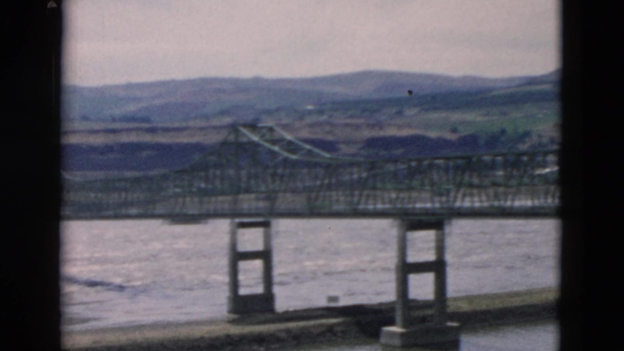 Steel bridge and river landscape recorded on 8mm film, framed by dark vertical gate edges and distant hills.