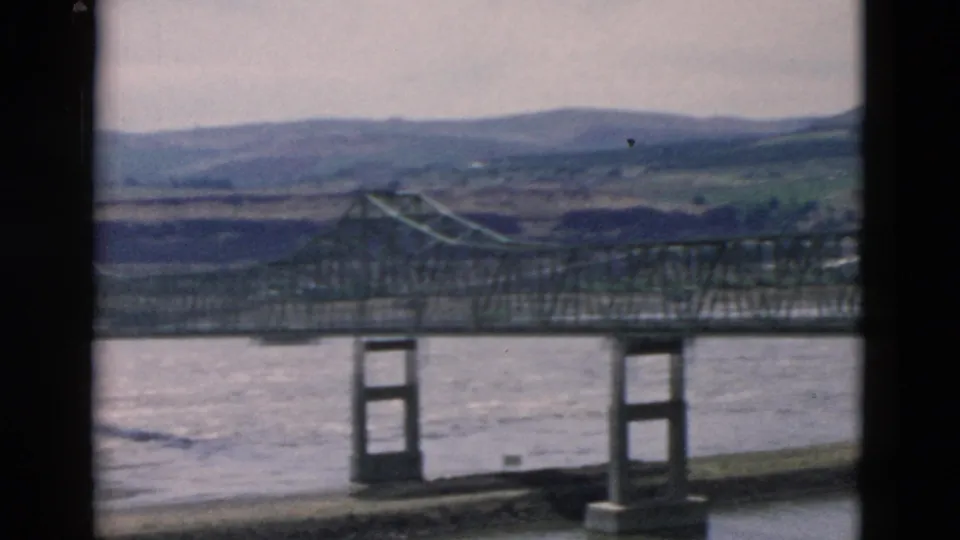 Steel bridge and river landscape recorded on 8mm film, framed by dark vertical gate edges and distant hills.