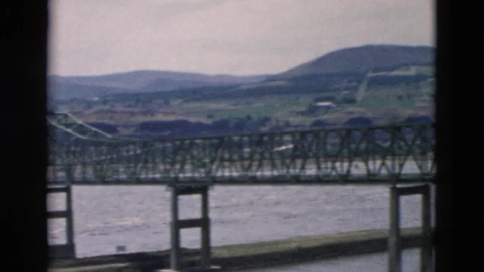 Vintage home-movie frame showing the steel truss bridge from a closer passing angle above the river and rural embankment.