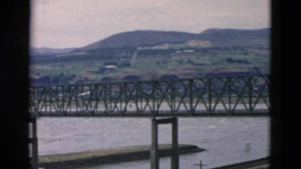 Steel truss bridge spans the foreground while a river, rural fields, hillside road, and low mountains appear beyond.