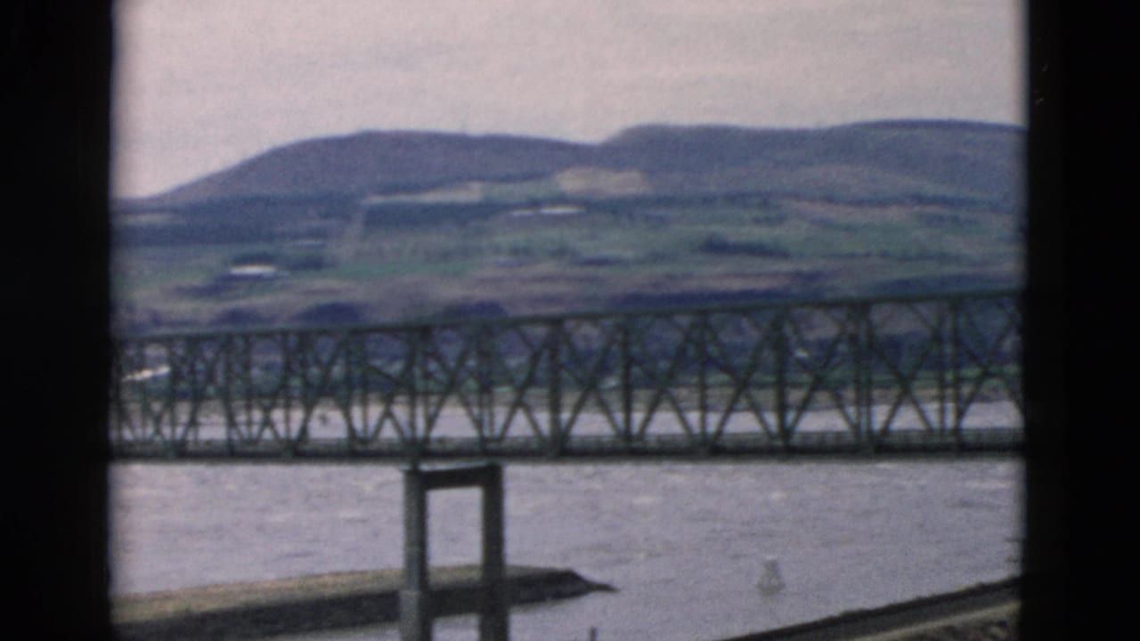 Iron truss bridge crossing a river with low hills in the background.