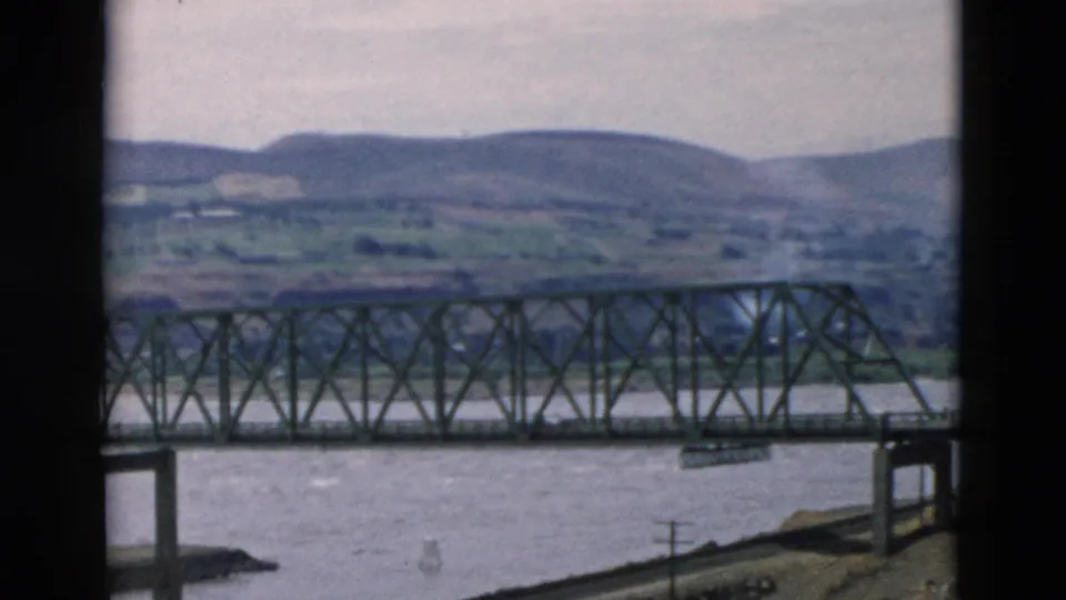 Iron truss bridge over water with rural hills and a hazy sky, seen through dark frame borders on 8mm film.