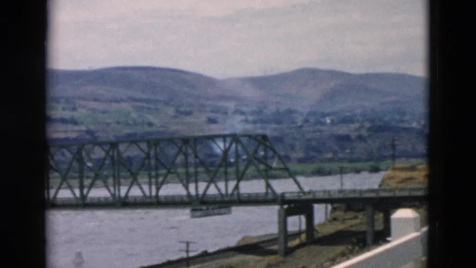 Rural river crossing with an iron bridge, low mountain backdrop, roadside edge, utility pole, and white fence in the foreground.