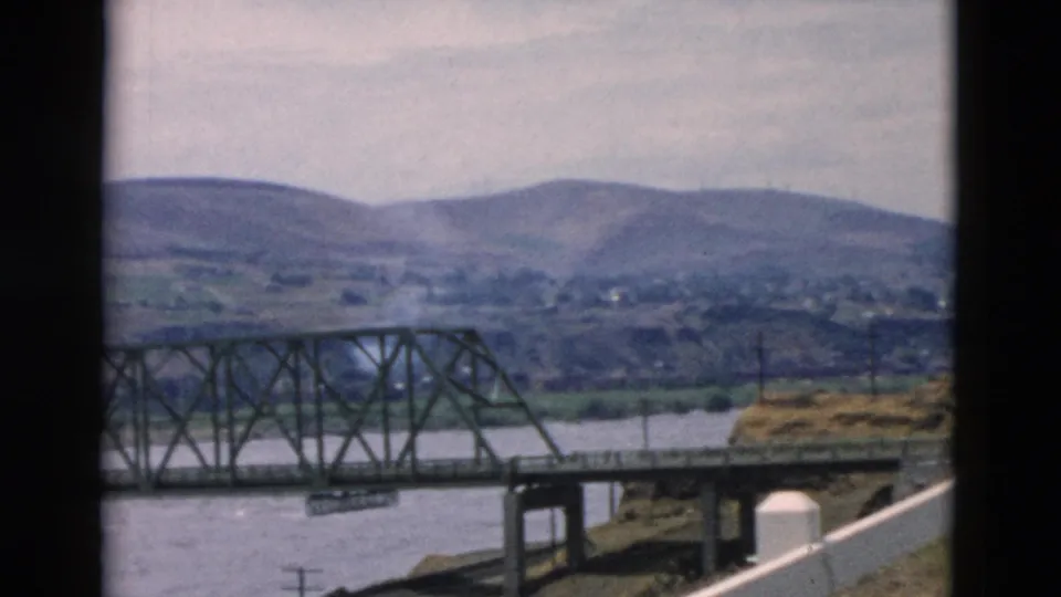 Final 8mm frame shows the bridge partly cropped, with river water, a roadside utility pole, white fence, and low hills beyond.