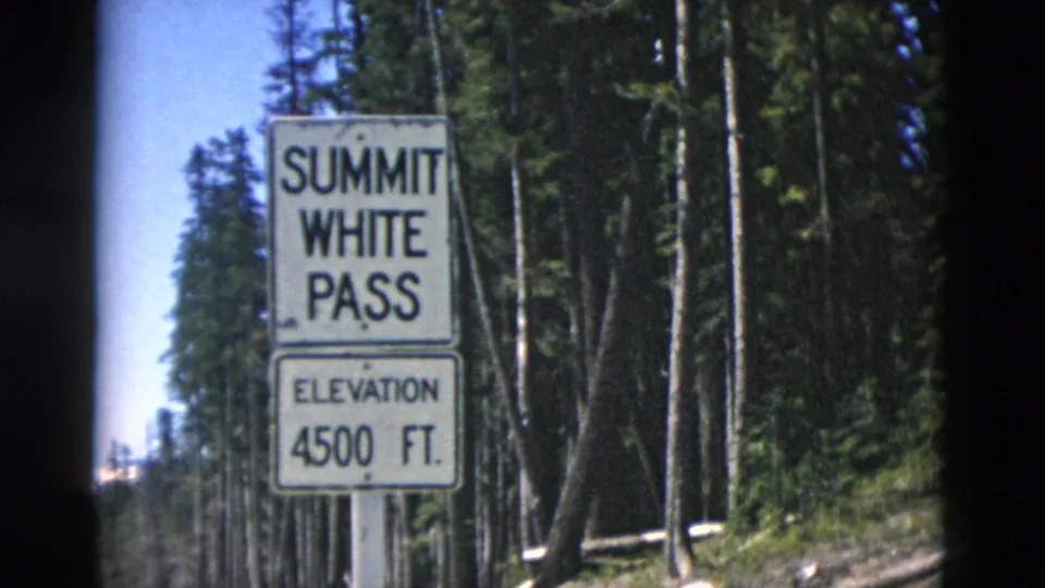 1958 8mm frame of the Summit White Pass elevation sign at 4500 ft, framed by dark conifers and roadside vegetation.
