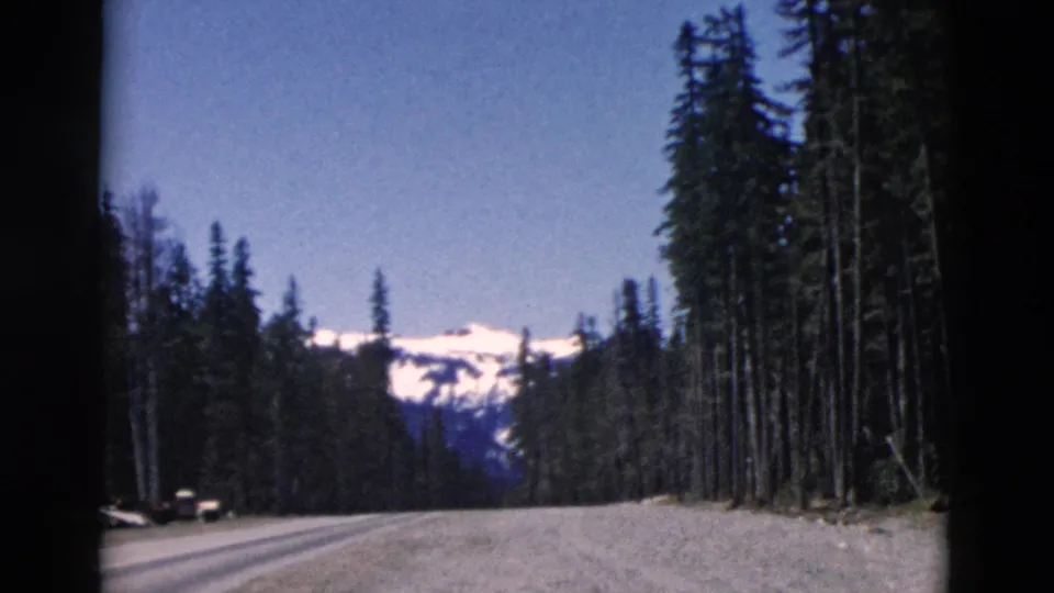 A handheld 8mm frame shows a forested mountain road with a small vehicle near the shoulder and snowy peaks beyond.