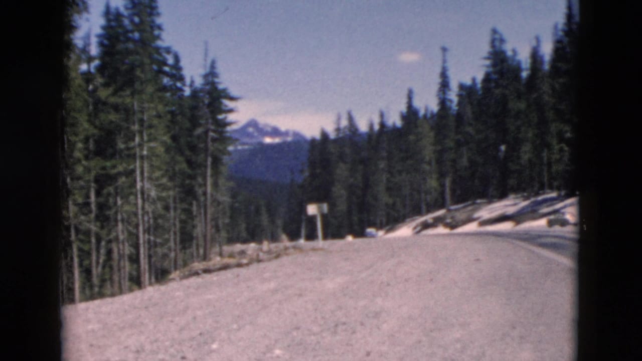 Wide road shoulder and pine forest with snowbanks and distant mountain peaks.
