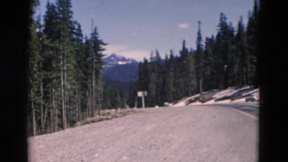 1958 Montana 8mm frame looking along a pale mountain road bordered by pines, snow patches, and distant peaks.