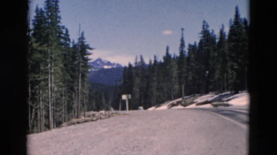 1958 Montana 8mm frame of a mountain road bending through dense pines, with snow on the roadside and peaks beyond.