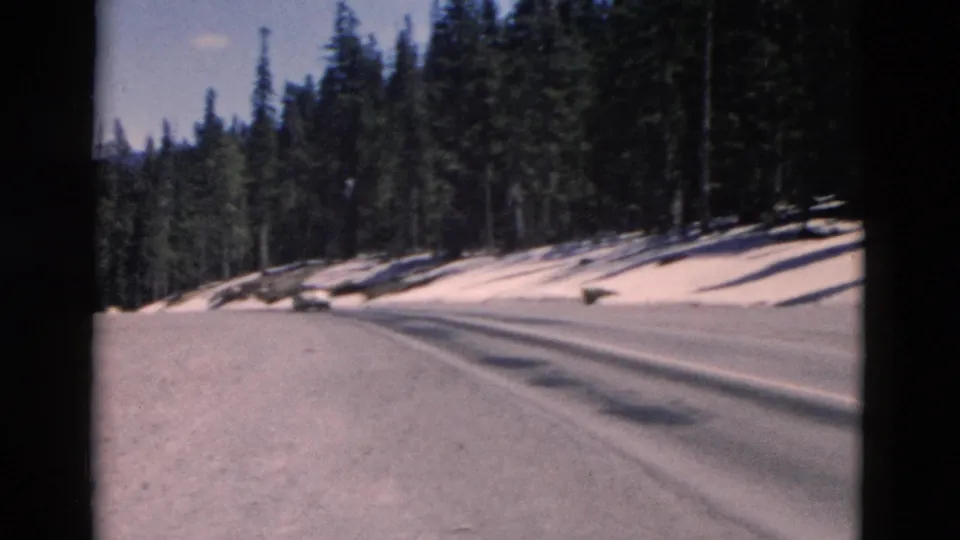 1958 Montana 8mm frame showing a snow-lined mountain road through pine forest, with long shadows across the pavement.