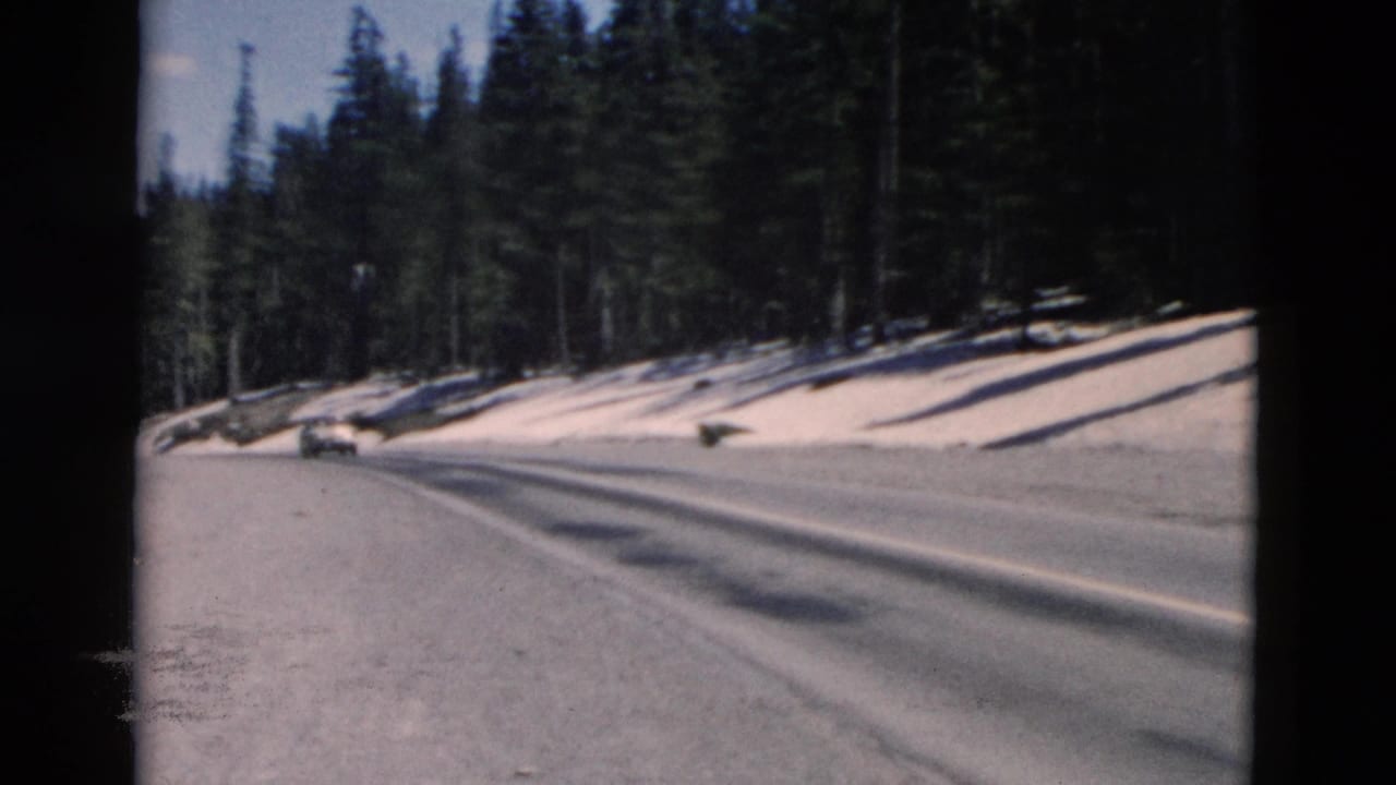 Curving mountain road beside evergreens and patches of snow, with a distant car visible.