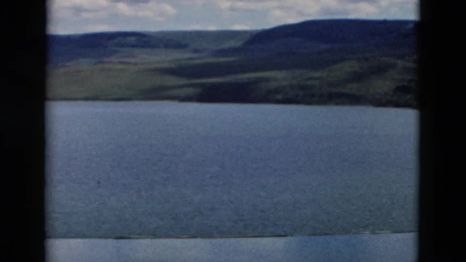 1958 Montana 8mm frame looking across a mountain lake toward rolling green hills under broken cloud cover.