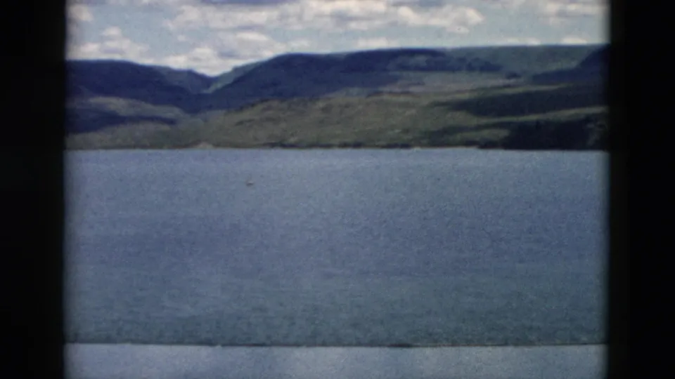 1958 Montana 8mm frame showing a wide mountain lake view with sloping hills and shoreline beneath a cloudy sky.