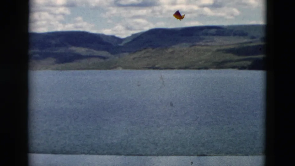 1958 Montana 8mm frame showing open lake water, distant green hills, dark mountain ridges, and cloudy sky through film gate edges.