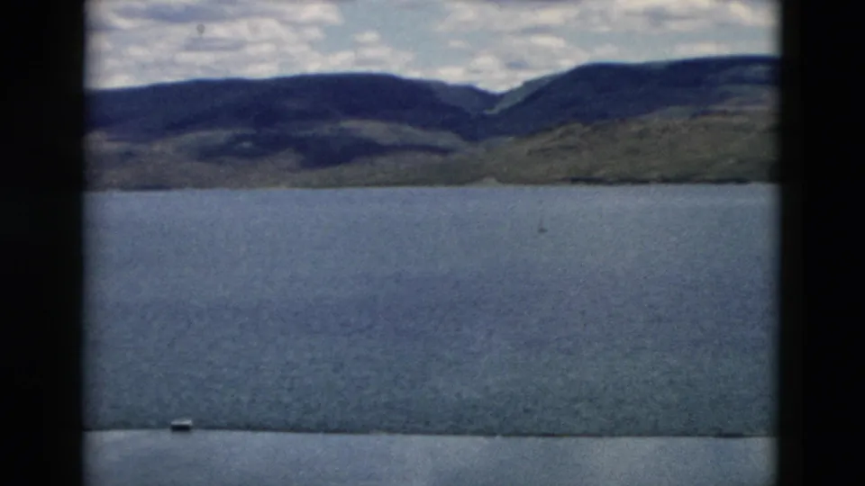 1958 Montana home-movie frame with a wide lake foreground, layered hills beyond, dark ridgeline, and visible 8mm frame borders.