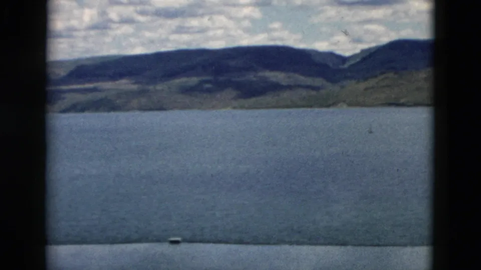 1958 Montana 8mm still showing lake water, distant hills, a mountain ridge, cloudy sky, and a dark strip from the scanned film edge.