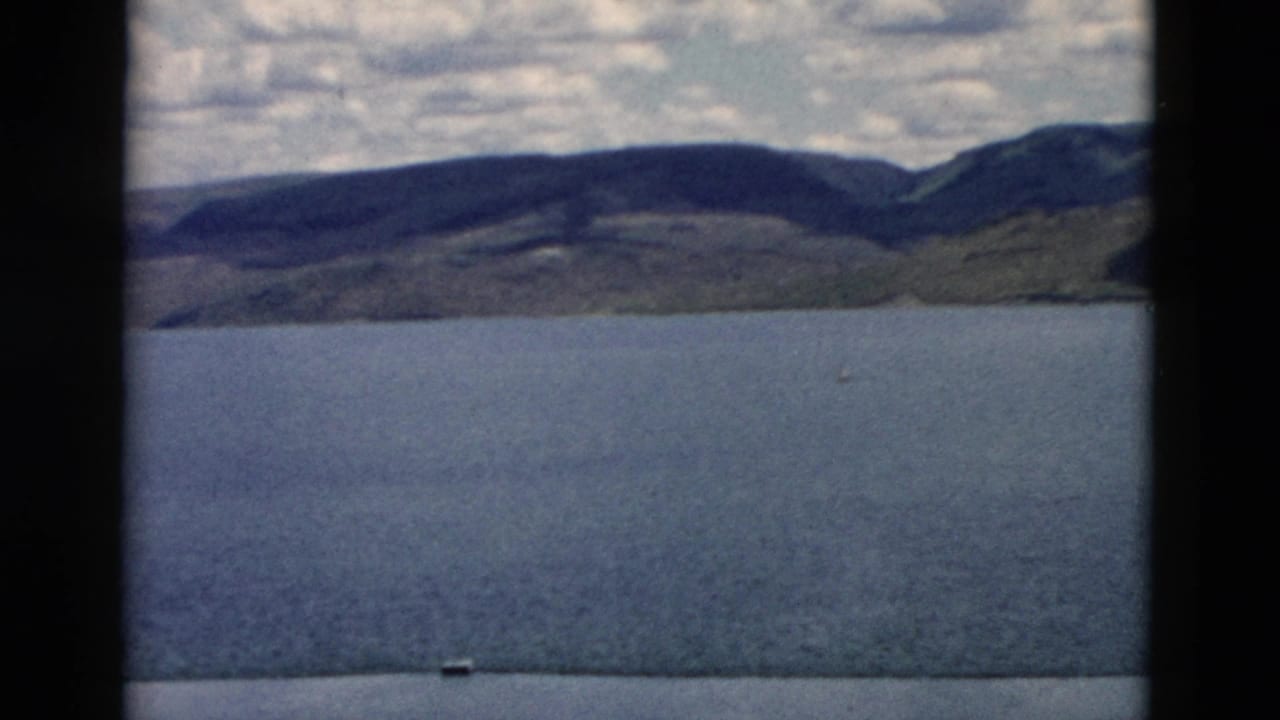 1958 Montana 8mm frame showing blue water, low distant hills, darker ridges, and cloud cover, with a tiny boat-like speck near the lower edge.