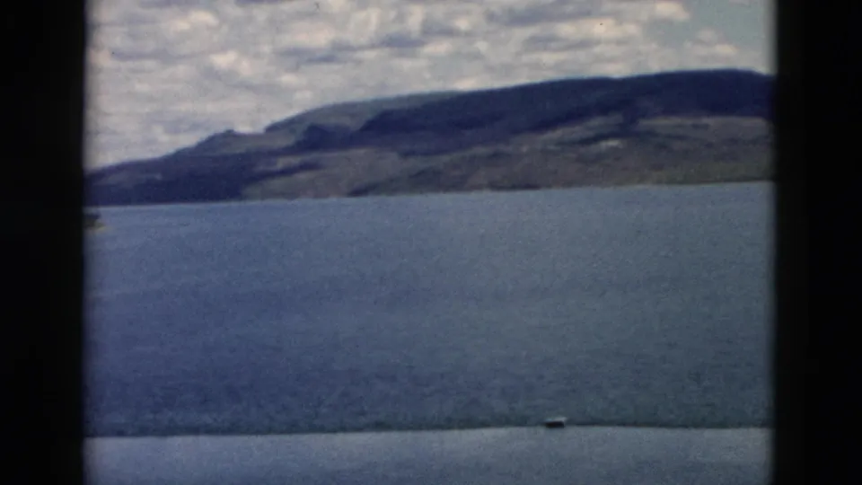 1958 Montana 8mm frame looking across blue water toward a long low hillside and darker mountain ridge under clouds.