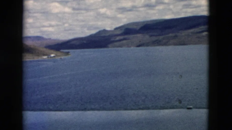 1958 Montana 8mm frame showing water, a curving shoreline, rolling hills, and a small light detail near the far shore.