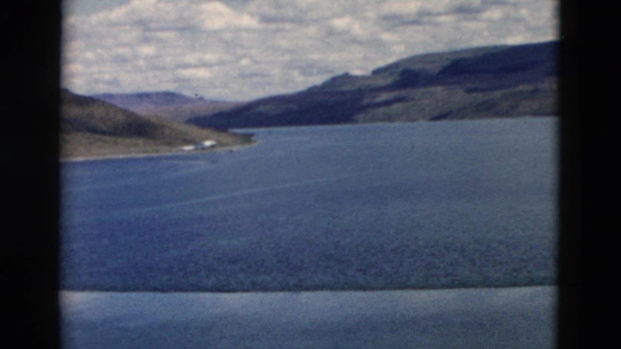 1958 Montana 8mm frame showing a calm blue lake, dry hills, cloudy sky, and dark window-like borders.