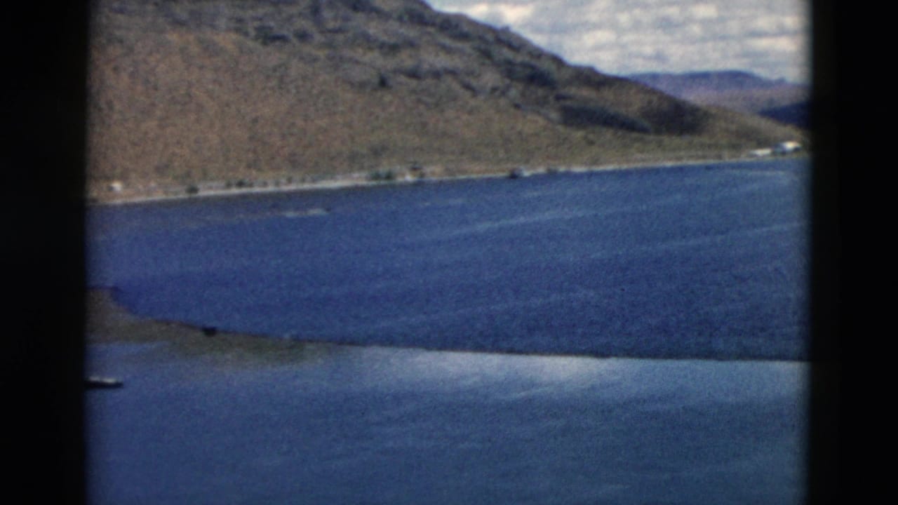 1958 Montana lake shoreline with brown hills, blue water, distant structures, and dark framing from the filming position.