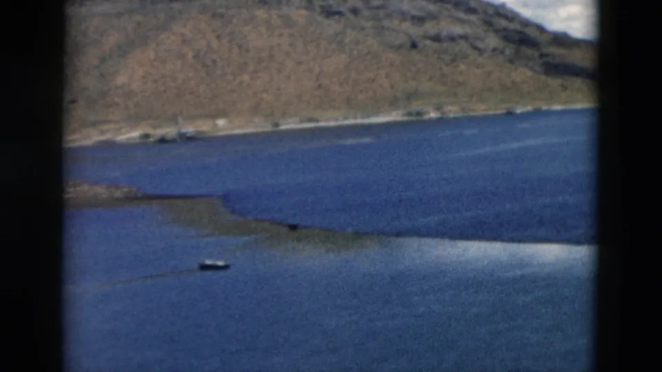 1958 Montana 8mm view of a small boat on blue water below arid hills and a low shoreline or island.