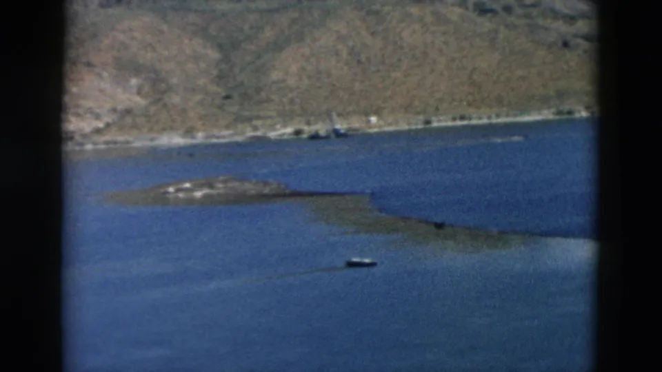 1958 Montana lake scene with a small boat, low island or spit, distant shoreline structures, and arid hills.