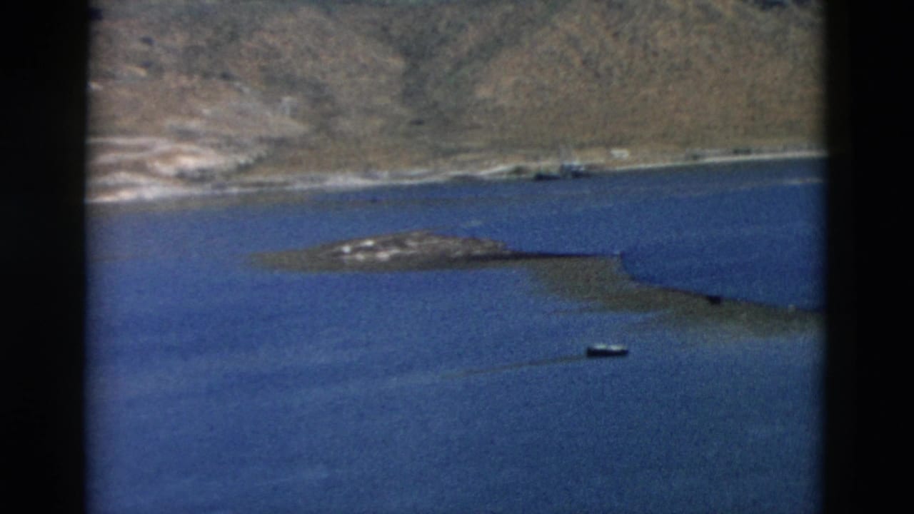 1958 Montana 8mm frame showing a blue lake, distant rocky hills, shoreline, and a small boat on calm water.
