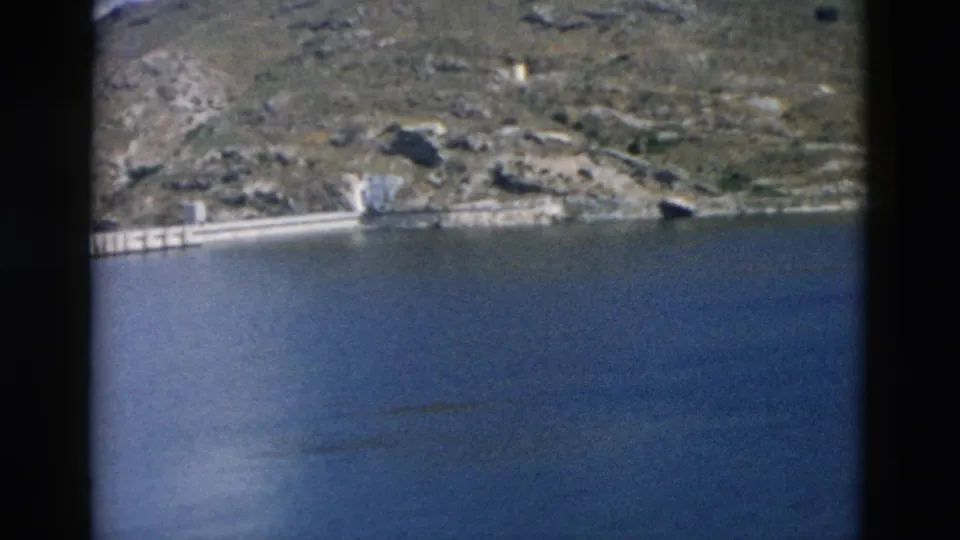 1958 Montana 8mm frame showing blue lake water below a rocky hillside and a concrete dam-like structure on the far bank.