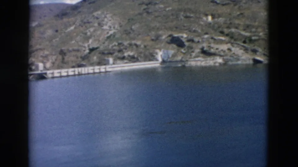 1958 Montana 8mm frame with blue lake water, a concrete dam-like shoreline structure, and dry rocky hills in the background.