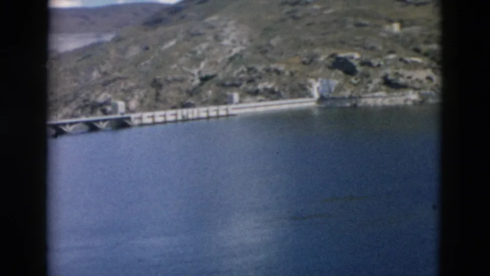 1958 Montana 8mm frame showing a long concrete bridge or dam-like span over blue water beside rugged hills.