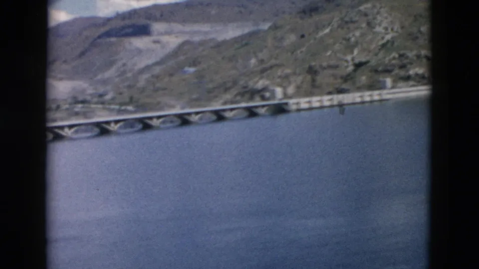 Soft 8mm frame of a long concrete crossing over water, with Montana hills and sparse structures in the background.