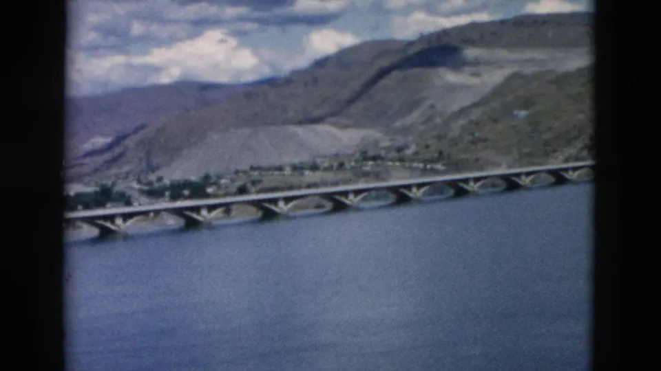 1958 Montana 8mm frame showing the concrete bridge from farther along the pan, with mountains, sky, and water visible.