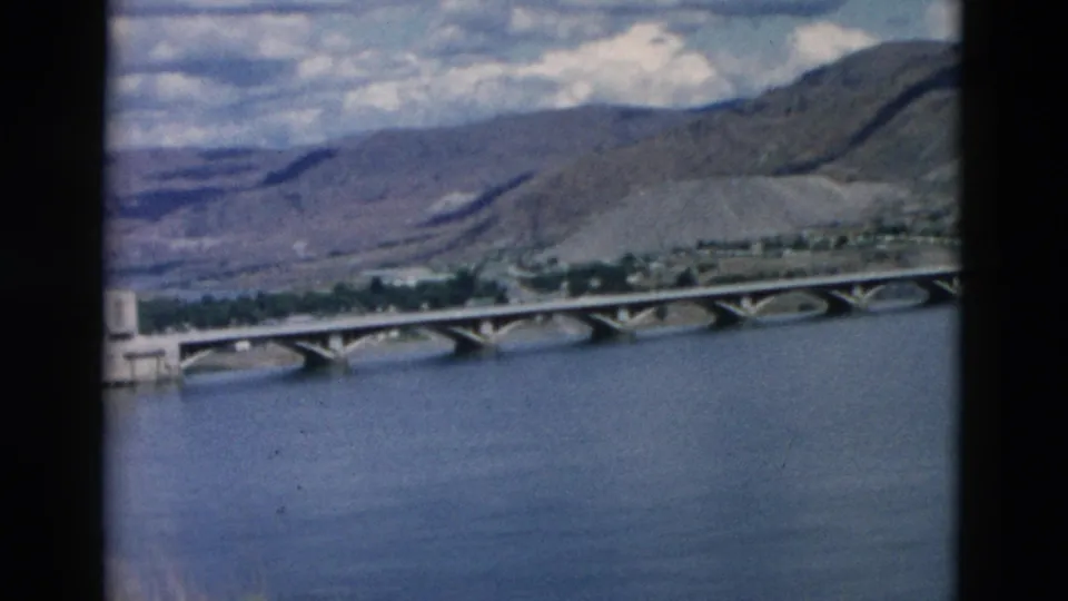 1958 Montana 8mm view of an arched bridge crossing a river, with shoreline trees and mountain ridges in the background.