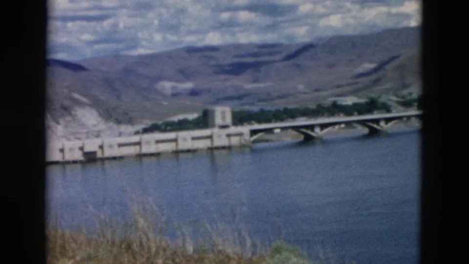 1958 Montana 8mm view of a riverside concrete dam structure with a bridge, dry grasses, water, and mountain landscape.
