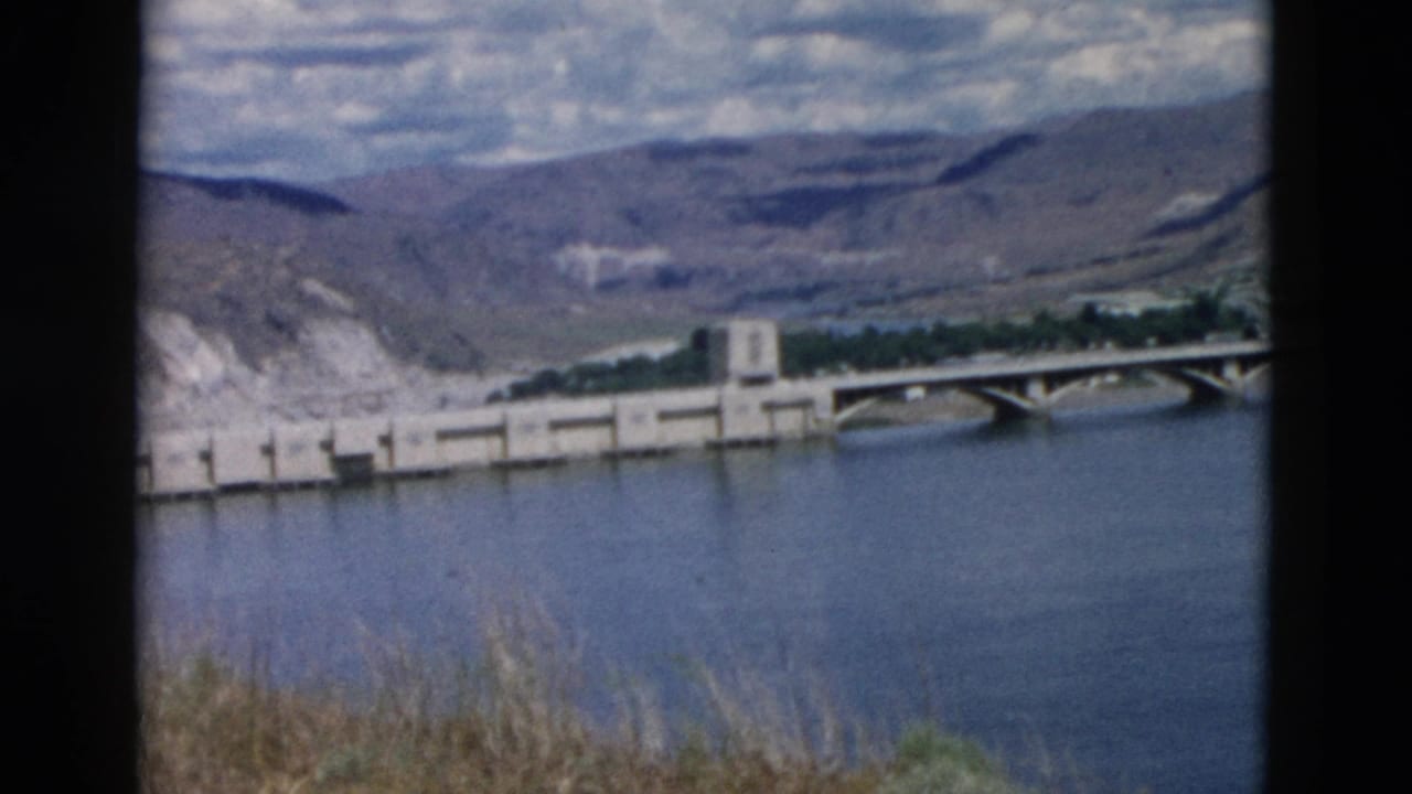 1958 Montana 8mm view of a concrete dam or bridge crossing a reservoir below dry hills and scattered clouds.
