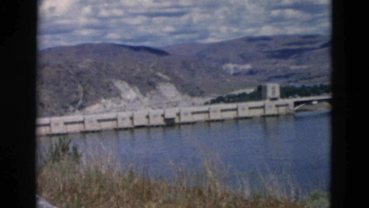 Dam or bridge structure crossing blue water with grasses in the foreground.