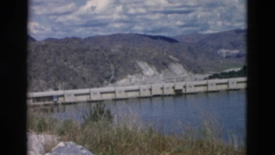 1958 Montana 8mm frame of a concrete dam or bridge beside still water, with rocks and grasses in the foreground.
