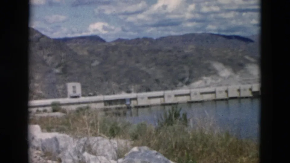 1958 Montana home-movie view of a reservoir crossing framed by foreground rocks, grasses, dry hills, and clouds.