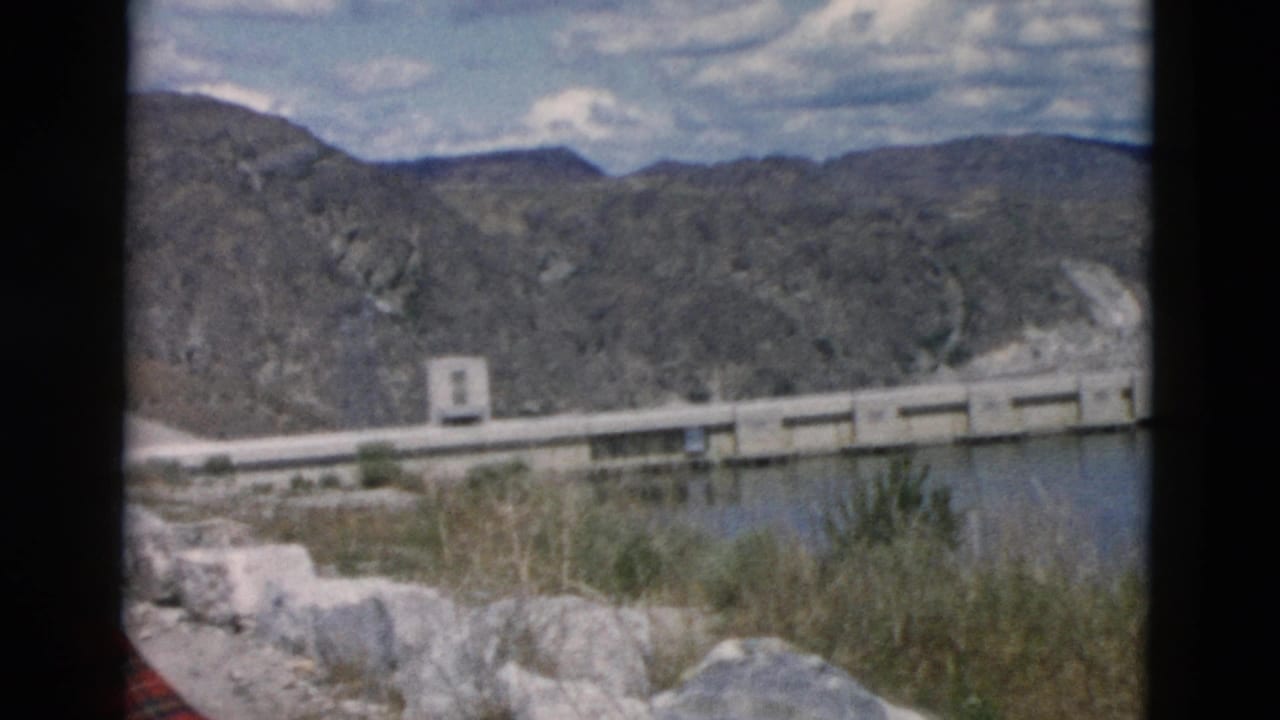 Concrete dam and reservoir below rugged mountains with rocks and brush in the foreground.