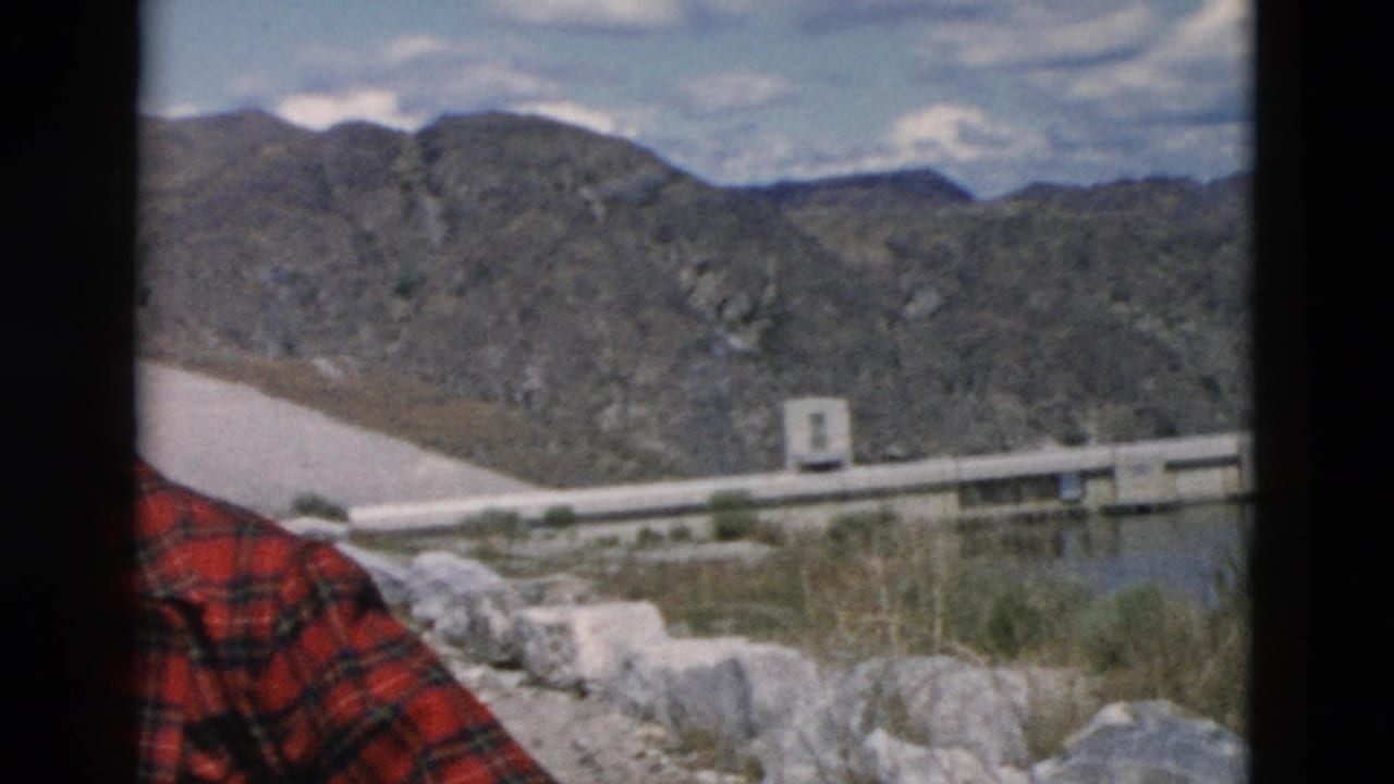 Part of a person in a red plaid coat stands before a dam, reservoir, and mountains.