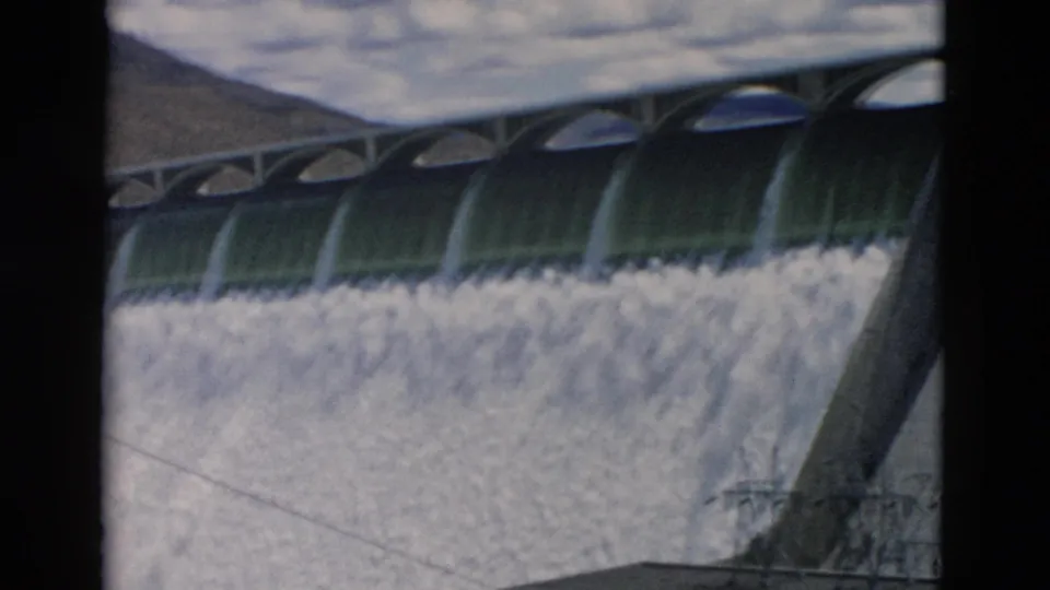 1958 Montana 8mm frame showing the dam spillway releasing water below arched concrete spans with distant mountain slopes.