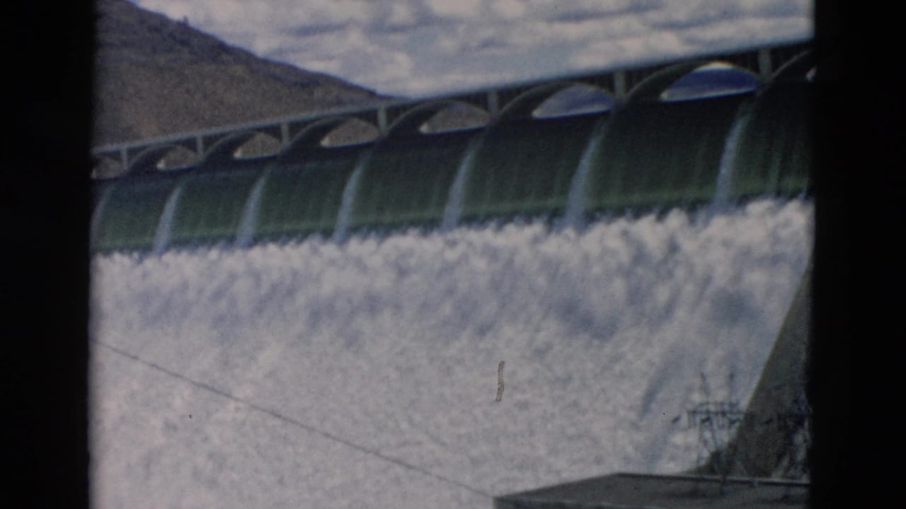1958 Montana home-movie frame showing arched dam gates above foamy water on a concrete spillway with mountains and cloudy sky behind.