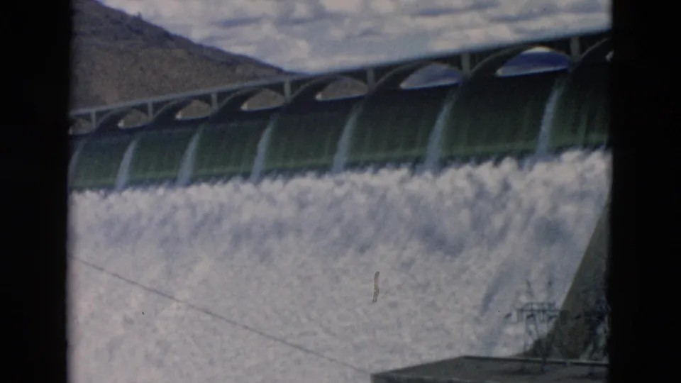 1958 Montana home-movie frame showing arched dam gates above foamy water on a concrete spillway with mountains and cloudy sky behind.