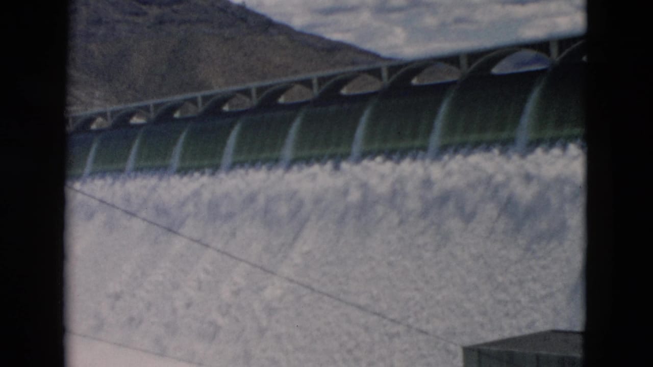 1958 Montana dam footage frame with arched spillway openings, foamy cascading water, a diagonal cable or line, and dark film borders.