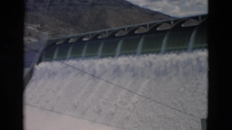 1958 Montana home-movie view of foamy water rushing down a concrete dam face beneath arched spillway gates.