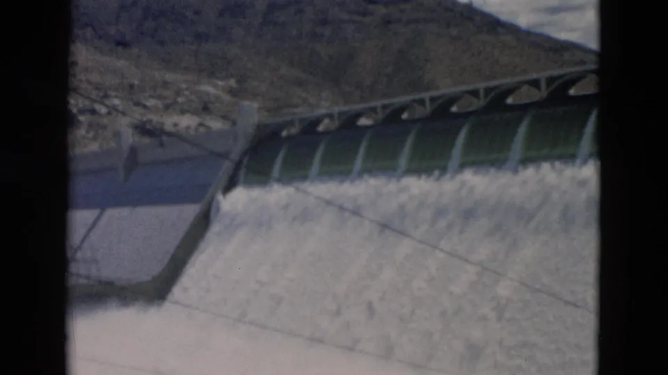 1958 Montana frame showing the dam from a slightly wider angle, with foamy spillway water, arched gates, mountain terrain, and film gate edges.