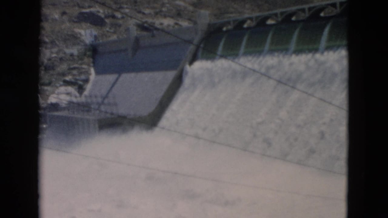 1958 Montana home-movie frame with green dam gates above a wide sheet of released water on a concrete face.